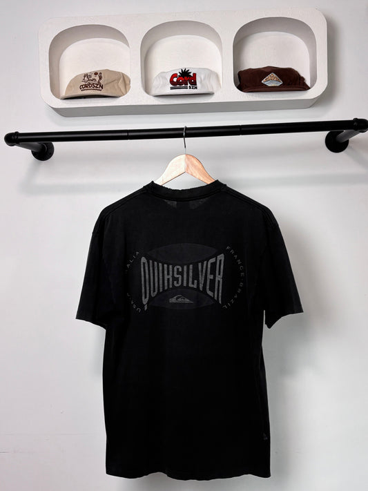 Black Quiksilver t-shirt hanging on a rack with shelves displaying hats in the background.