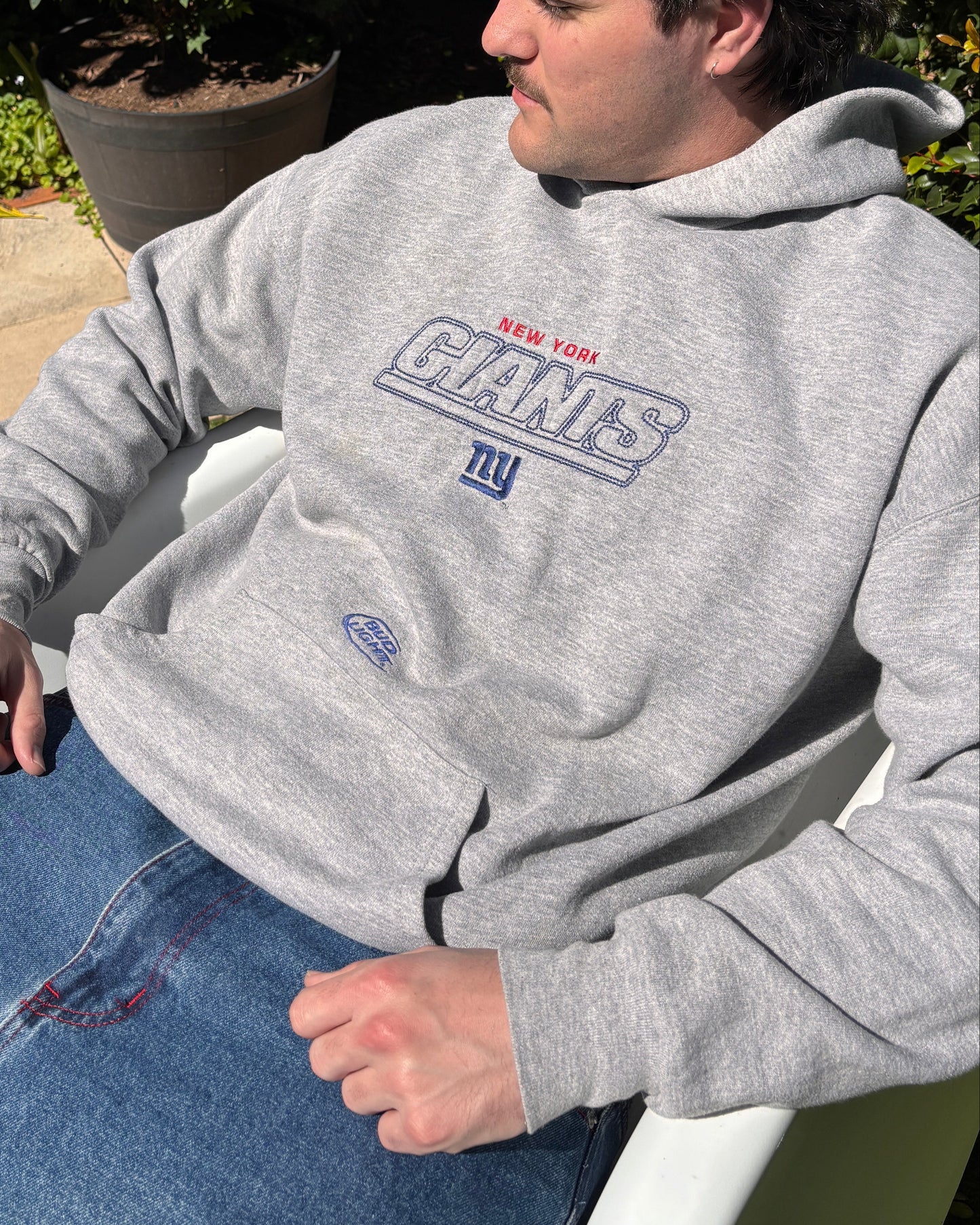 A man sitting outdoors in a white chair wearing a heather gray New York Giants hoodie and blue jeans, with garden plants and grass in the background.
